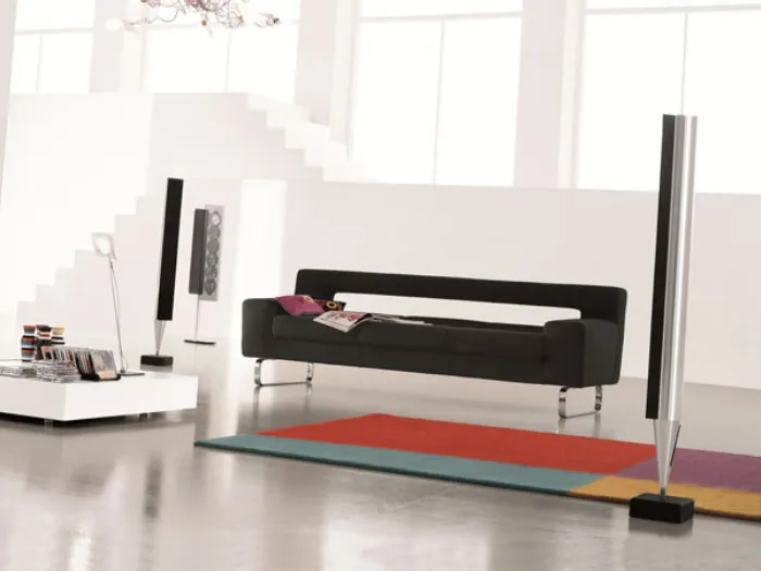 divano a tre posti nero con gambe in alluminio inserito in spazio living bianco con tappeto geometrico multicolore e casse verticali