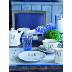 servizio da tè con teiera bianco con decorazione di lavanda, bicchiere in vetro blu sfaccettato, fiori blu e porta candela trasparente blu su tavolo in legno
