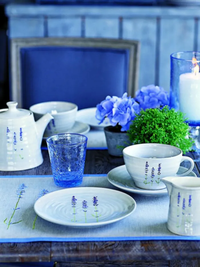 servizio da tè con teiera bianco con decorazione di lavanda, bicchiere in vetro blu sfaccettato, fiori blu e porta candela trasparente blu su tavolo in legno