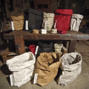 tre sacchi in primo piano panna, nocciola e corda, tavolo in legno grezzo con altri sacchi anche rossi e neri