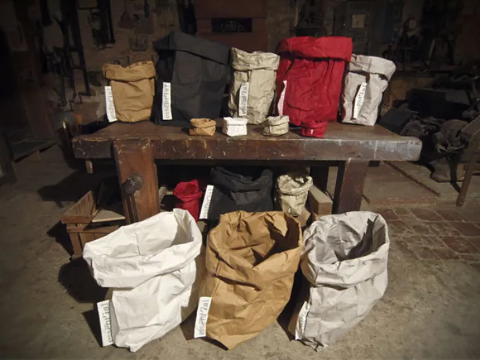 tre sacchi in primo piano panna, nocciola e corda, tavolo in legno grezzo con altri sacchi anche rossi e neri