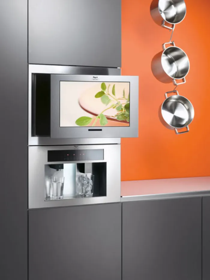 televisore incassato in mobile cucina grigio, tre pentole sospese a catena con parete di sfondo arancione