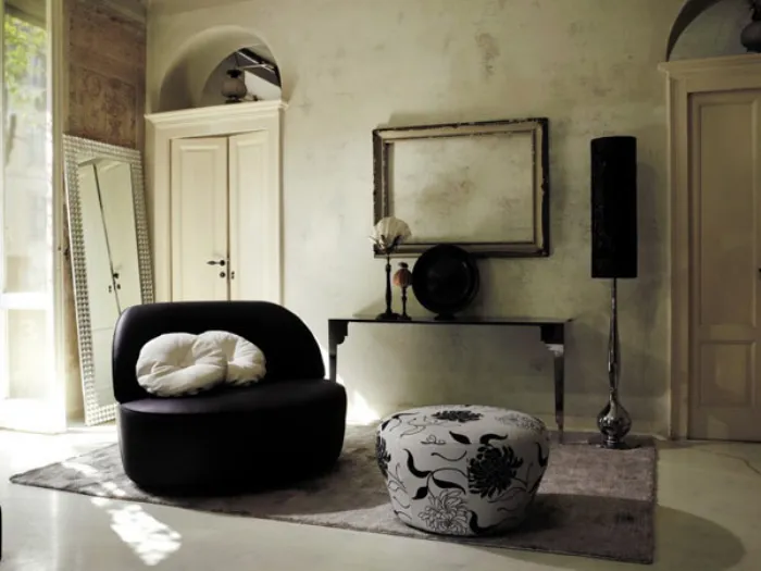 soggiorno con poltrona a due posti nera e cuscini bianchi, pouf bianco con grafiche floreali nere, tappeto, cornice vuota appesa, cassa con sostegno decòr
