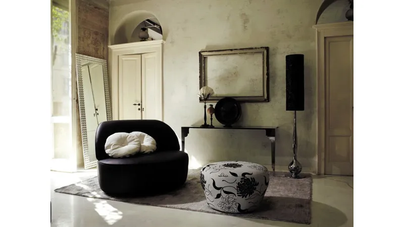 soggiorno con poltrona a due posti nera e cuscini bianchi, pouf bianco con grafiche floreali nere, tappeto, cornice vuota appesa, cassa con sostegno decòr