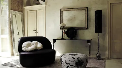 soggiorno con poltrona a due posti nera e cuscini bianchi, pouf bianco con grafiche floreali nere, tappeto, cornice vuota appesa, cassa con sostegno decòr