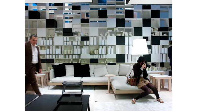 spazio espositivo con mobili zona living con libreria a parete bianca e nera, divano con chaise-longue in legno chiaro e tessili in ecrù e nero, due visitatori