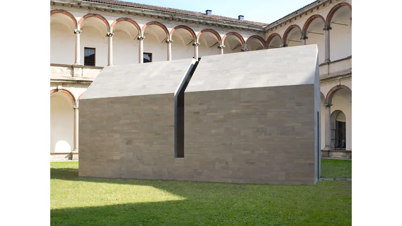 casa in pietra grigia con apertura verticale nel centro all'interno di chiostro con prato