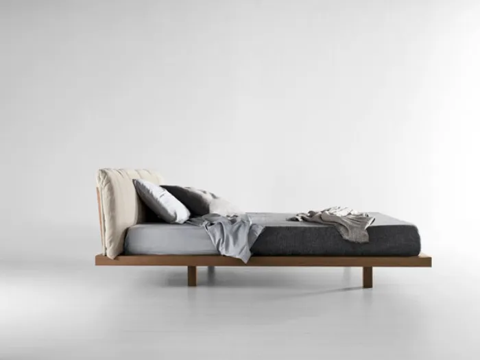 vista laterale di letto matrimoniale con struttura in legno, testiera di cuscini color panna e biancheria nei toni del grigio 