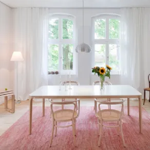 tavolo da pranzo con quattro sedie in legno chiaro, tre tavolini estraibili in legno chiaro, tappeto rosa e due finestre bianche con tende bianche