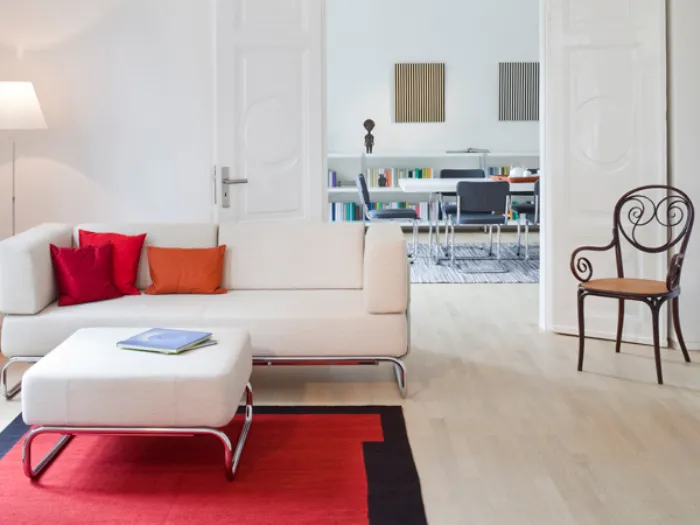 zona living con divano e pouf bianco con gambe in acciaio, cuscini e tappeto rossi, sedia in legno nero e seduta in legno naturale