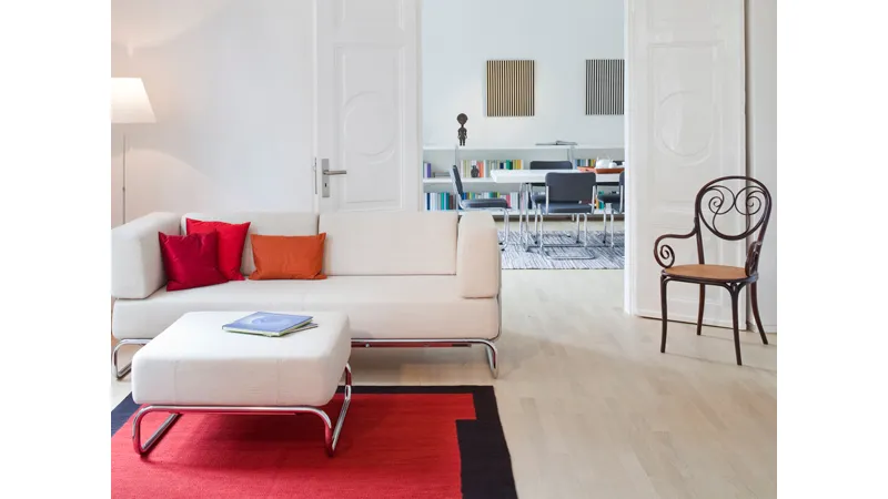 zona living con divano e pouf bianco con gambe in acciaio, cuscini e tappeto rossi, sedia in legno nero e seduta in legno naturale