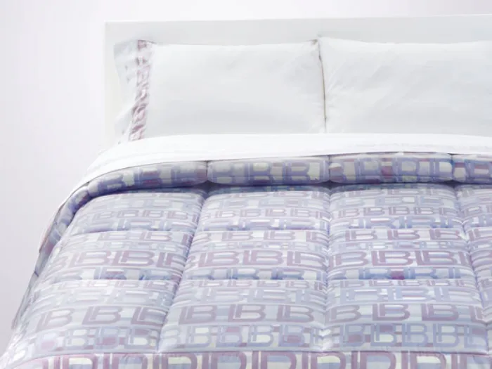 letto matrimoniale con biancheria bianca e copriletto con logo dai toni pastello lavanda e lilla